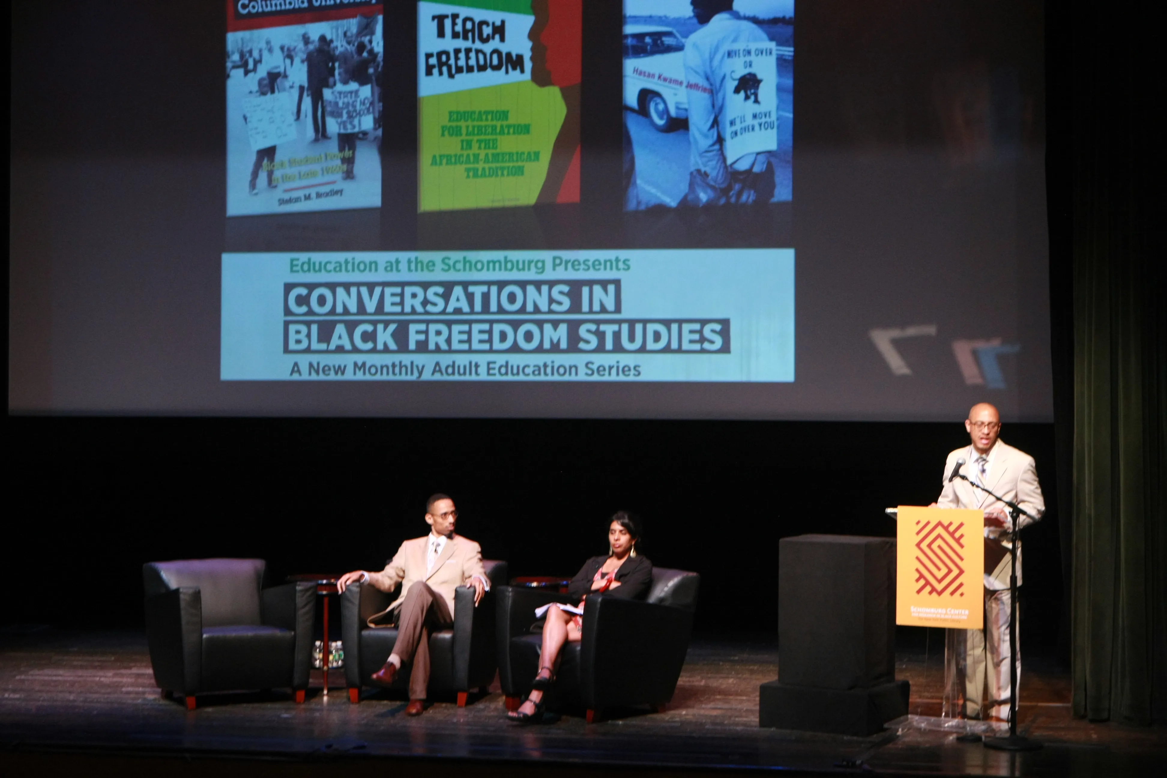 Two panelists seated on stage and a speaker at a lectern at the Schomburg Center, beneath a projected slide reading Conversations in Black Freedom Studies